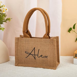 Op de afbeelding: Een rechthoekige bruine jute draagtas met bijpassende handvatten. De naam "Alex" is in zwart script op de voorkant van de tas geborduurd. De tas staat op een witte ondergrond.