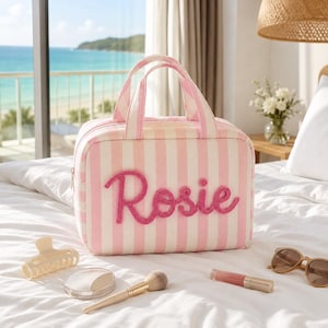 May include: A rectangular travel bag featuring vertical light pink and white stripes with the name Rosie embroidered in raised pink script on the front. The bag has two top handles and a zipper closure, accompanied by a hair clip, makeup brush, lipstick, and sunglasses.