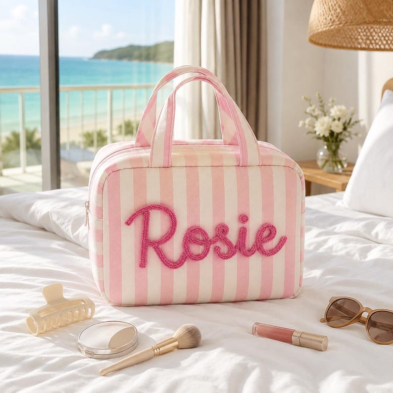 May include: A rectangular travel bag featuring vertical light pink and white stripes with the name Rosie embroidered in raised pink script on the front. The bag has two top handles and a zipper closure, accompanied by a hair clip, makeup brush, lipstick, and sunglasses.