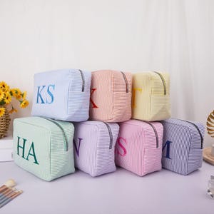 May include: A collection of personalized cosmetic bags in various pastel colors, including blue, orange, yellow, green, purple, pink, and navy. Each bag features embroidered initials and a zipper closure. The bags are striped and rectangular.