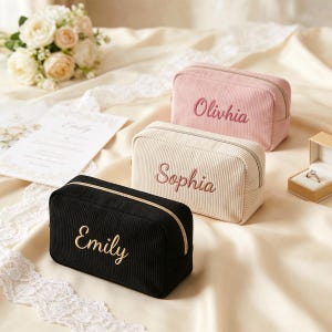 Pu&ograve; includere: Tre trousse per cosmetici in velluto a coste nere, rosa e crema, ciascuna con un nome ricamato. La borsa nera ha ricami dorati, mentre le borse rosa e crema hanno ricami rosa. Le borse sono rettangolari con chiusura a zip.