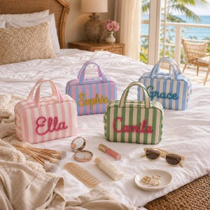 May include: Four rectangular striped cosmetic bags with top handles and embroidered names Ella, Sophie, Camila, and Grace. Accessories include makeup brushes, a compact mirror, a lip gloss, a lotion bottle, sunglasses, and a small dish with jewelry.