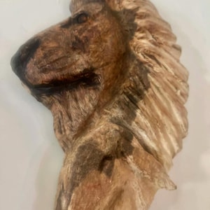 May include: A detailed, side profile of a lion's head sculpture. The sculpture is primarily brown with darker brown and tan accents, showcasing the mane and facial features. The lion's nose and eye are visible, adding depth to the artwork.