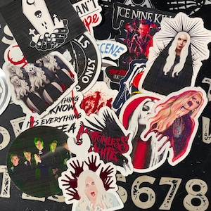 Puede incluir: Una colección de pegatinas con varios diseños, incluyendo un ataúd con una luna creciente, una banda con el texto "Motionless in White" y una mujer con una corona de espinas. Las pegatinas están sobre un fondo negro con un patrón blanco.