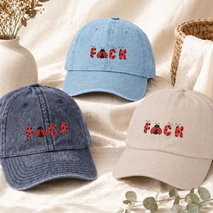 Könnte beinhalten: Drei Baseballkappen in Hellblau, Marineblau und Beige. Jede Kappe hat das Wort "FACK" in Rot mit einem Marienkäfer-Design gestickt. Die Kappen sind auf einer cremefarbenen Oberfläche angeordnet.