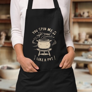 Op de afbeelding: Zwarte schort met de tekst "You spin me right round like a pot!" in een speels lettertype. Het ontwerp toont een pot op een standaard met muzieknoten. De schort heeft een zak en een nekband.