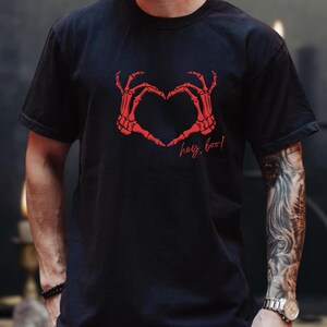 Peut inclure: T-shirt noir avec un motif de cœur formé de mains de squelette rouges et le texte "hey, boo!". Ce t-shirt décontracté est idéal pour Halloween ou les événements à thème.