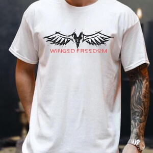 May include: White t-shirt featuring a black and red winged graphic and the text "Winged Freedom". The design is centered. The shirt has a crew neck and short sleeves. A casual style.