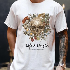 May include: White t-shirt with a skull design encircled by flowers, mushrooms, and a dragonfly. The text "Life & Death" is printed below the graphic, with "A Beautiful Balance" underneath. The design is in a watercolour style.