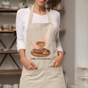 May include: Beige apron with a large front pocket and neck strap. The apron features an illustration of hands shaping a clay pot, with the text "Made with Mud & Love" below the image. The apron is worn by a person.