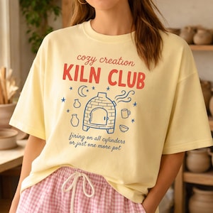 Op de afbeelding: Een lichtgeel t-shirt met de tekst "cozy creation KILN CLUB" in rood en zwart. Het shirt heeft een ovenafbeelding en de zin "firing on all cylinders or just one more pot". De persoon draagt een roze en witte geruite short.