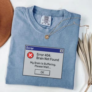 以下が含まれることがあります： コンピューターのエラーメッセージを模倣したグラフィックが特徴の、ライトブルーのComfort Colors Tシャツ。「Error 404: Brain Not Found. My Brain Is Buffering. Please Wait...」と書かれています。ペンダント付きのゴールドネックレスがシャツの上にかけられています。