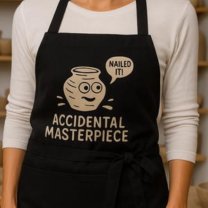 Pode incluir: Avental preto com um design de cerâmica extravagante e o texto "ACCIDENTAL MASTERPIECE". O design inclui um pote sorridente com o texto "NAILED IT!" numa bolha de fala. O avental tem um bolso frontal e uma gravata.