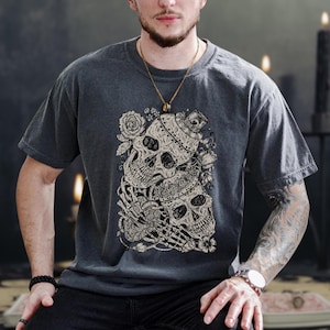 May include: Charcoal grey t-shirt with a detailed graphic of two skulls entwined with roses and skeletal hands. The skulls are decorated with ornate details. The shirt has a classic crew neck and short sleeves.