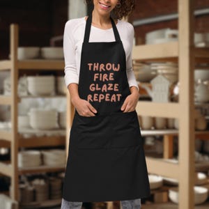 Puede incluir: Delantal negro con el texto "THROW FIRE GLAZE REPEAT" en letras rosas. El delantal tiene un peto, dos bolsillos delanteros y una correa para el cuello. El fondo muestra estanterías con cerámica.