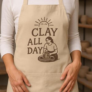 May include: Beige apron with the text "CLAY ALL DAY" and an illustration of a person working on a pottery wheel. The apron has a front pocket and is worn over a white long-sleeved shirt.
