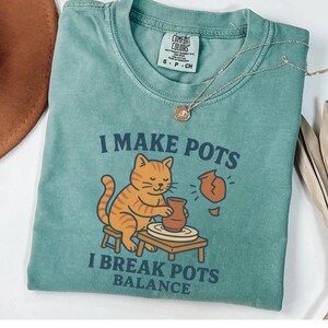 Może przedstawiać: Turkusowa koszulka Comfort Colors z napisem "I MAKE POTS I BREAK POTS BALANCE" oraz ilustracją pomarańczowego kota pracującego na kole garncarskim. Złoty naszyjnik z wisiorkiem jest zarzucony na koszulkę.