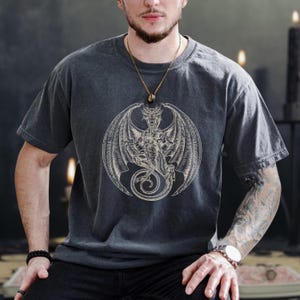 Peut inclure: T-shirt gris foncé avec un motif de dragon blanc détaillé dans un cercle. Le dragon a des ailes déployées et une longue queue enroulée. Le t-shirt est à manches courtes et à col rond.