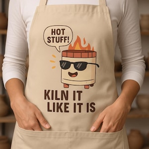 Może przedstawiać: Beżowy fartuch z kreskówkowym wzorem pieca w okularach przeciwsłonecznych i płomieniach. Tekst brzmi "HOT STUFF!" w dymku i "KILN IT LIKE IT IS" poniżej. Fartuch ma dwie przednie kieszenie.