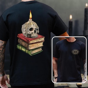 Peut inclure: T-shirt noir avec une illustration détaillée d'une bougie en forme de crâne sur une pile de livres. La flamme de la bougie est jaune et orange. Une petite image montre un motif d'œil doré sur le devant du t-shirt.