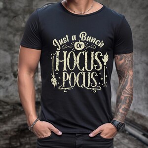 May include: Black t-shirt featuring the text "Just a Bunch of Hocus Pocus" in a vintage font. The design includes a witch's broom, a pumpkin, and stars. The shirt has a crew neck and short sleeves.