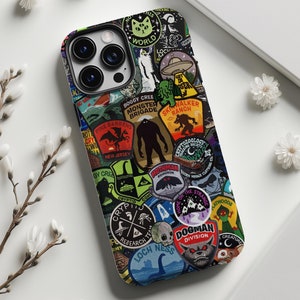 Könnte beinhalten: Eine schwarze iPhone-Hülle mit einem bunten Collage aus Aufklebern, die verschiedene Kryptide, Monster und Fabelwesen zeigen. Die Aufkleber enthalten Text wie "Loch Ness", "Dogman Division", "Monster Brigade" und "Cryptozoology Research Team".