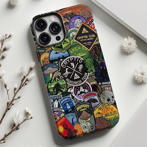 以下が含まれることがあります： カラフルなステッカーのコラージュが施された黒いiPhoneケース。ステッカーには、クリプティッド、モンスター、陰謀論などが描かれており、「クリプティッドクラブ」、「エリア51」、「ビッグフットアーミー」、「クリプティッド野生生物を保護！」などのテキストが含まれています。