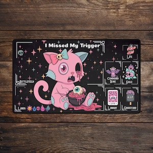 May include: A black rectangular playmat featuring the text I Missed My Trigger. The design displays a pink cartoon cat with a skull bow, holding a bowl of brains and an eyeball. It includes labeled zones for Battlefield, Lands, Command Zone, Exile, Library, and Graveyard.
