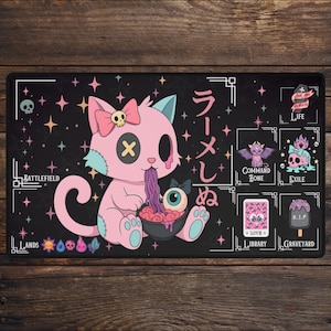 May include: Black rectangular playmat featuring a pink cat with a skull bow, eating ramen. The mat has Japanese text and various game-related icons, including a skull, bat, and ice cream. The background is black with star accents.