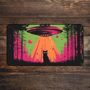 Puede incluir: Alfombra rectangular negra con una ilustración retro de un OVNI sobre un bosque. El diseño presenta un gato negro con ojos amarillos, un haz de luz naranja y árboles rosas y verdes.