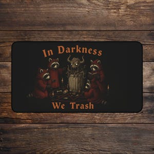 May include: Black rectangular mat with the text "In Darkness We Trash". The mat features an illustration of raccoons in red hooded robes gathered around a trash can with a horned creature. Candles and bones are also present.