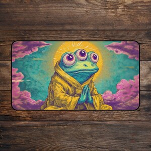 Puede incluir: Alfombra rectangular con una rana de dibujos animados de tres ojos, vestida con una túnica amarilla y con las manos juntas en oración. La rana está situada sobre un fondo de sol amarillo, cielo azul y nubes rosas.