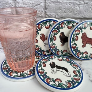 Puede incluir: Un juego de posavasos de cerámica con ilustraciones de Cavalier King Charles Spaniel. Cada posavasos tiene un fondo blanco, bordes florales y un borde azul. Un vaso rosa se encuentra sobre uno de los posavasos.