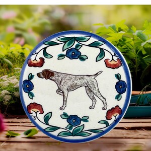 May include: Round ceramic coaster featuring a German Shorthaired Pointer dog illustration. The coaster has a white background with a blue border and is decorated with blue and red floral accents. The dog is brown and white.