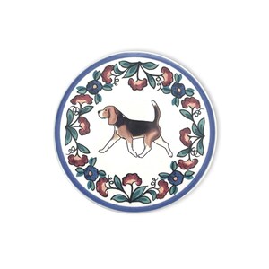 May include: Circular ceramic coaster with a beagle dog illustration. The coaster has a white background, a blue border, and a floral wreath design in blue, red, and green. The beagle is depicted in brown, black, and white.