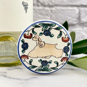 May include: A round, ceramic coaster with a cream-colored Cocker Spaniel illustration. The coaster is decorated with blue and red floral accents and a blue border. The background is white.