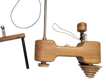 May include: Two wooden spinning tools are displayed against a white background. One is a small, rectangular frame with black legs. The other is a larger, more complex tool with a wooden body, metal components, and a conical base. A green rubber band is also present.