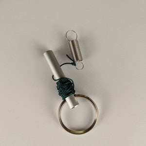 May include: A silver metal key ring with a silver metal rod and a green cord wrapped around it. The rod is attached to a silver metal spring.