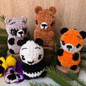 Pode incluir: Quatro animais de pelúcia de crochê: um panda, um urso, um guaxinim e uma raposa. Os animais estão todos sentados em uma superfície de madeira e são cercados por galhos de pinheiro verdes.