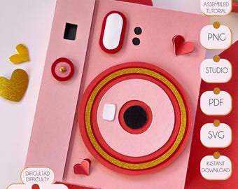 May include: Pink and red papercraft camera with gold glitter accents. The camera is a 3D model with a pop-up lens and a heart on the side. The image includes the text "DIFICULTAD DIFFICULTY" and "BAJA MEDIA ALTA LOW HALF HIGH".