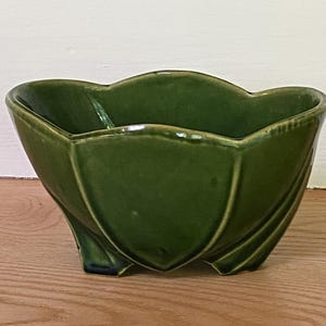 May include: A glossy olive green ceramic bowl with a scalloped rim and a petal-like exterior design. The bowl features three small feet at the base and a smooth, reflective finish that highlights the curved, organic shape of the vessel.