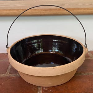 May include: A round, shallow ceramic bowl with a matte, light tan exterior and a glossy, dark brown glazed interior. The bowl features a wide, flat rim and is equipped with a thin, arched metal wire handle attached to two small side lugs.
