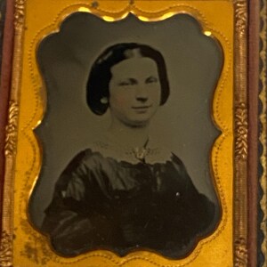 May include: Antique portrait of a woman in a decorative frame. The image is a sepia tone, showing a woman with dark hair and a dark dress. The frame is gold and brown, with ornate detailing.