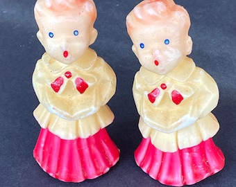 May include: Two vintage candle figurines of children wearing yellow robes and red skirts. The candles have blue eyes and red painted details.