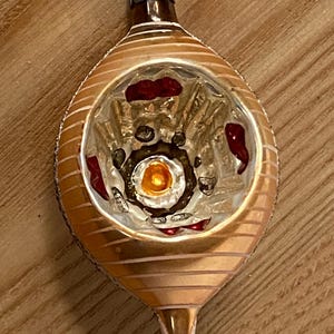 May include: Vintage Christmas ornament with a gold and brown color scheme. The teardrop-shaped glass ornament features a striped pattern and interior details in red and gold. A small metal loop is at the top for hanging.
