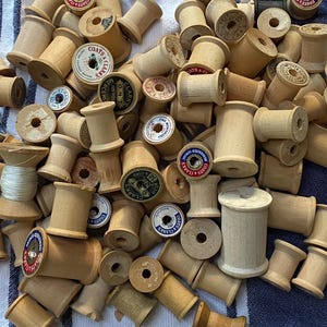 May include: A collection of numerous empty wooden sewing thread spools of varying sizes and shades of light brown. Many spools feature circular paper labels on the ends with printed text including brand names like Coats and Clark's.