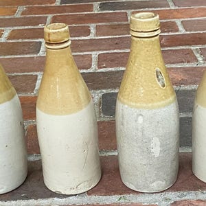 May include: Four vintage ceramic bottles with a light brown top and a white bottom. The bottles are arranged in a row on a brick surface.