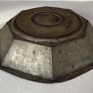 May include: A vintage, octagonal metal bowl with a weathered, tarnished appearance. The bowl features a central, raised circular area and a series of angled sides. The metal has a mottled, aged patina.