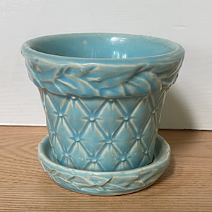 May include: A light blue ceramic flower pot with a matching saucer. The pot has a diamond pattern and leaf design around the rim. The pot and saucer are made of ceramic.