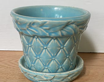 Pode incluir: Um vaso de flores de cerâmica azul claro com um pires a condizer. O vaso tem um padrão de diamante e um design de folha ao redor da borda. O vaso e o pires são de cerâmica.